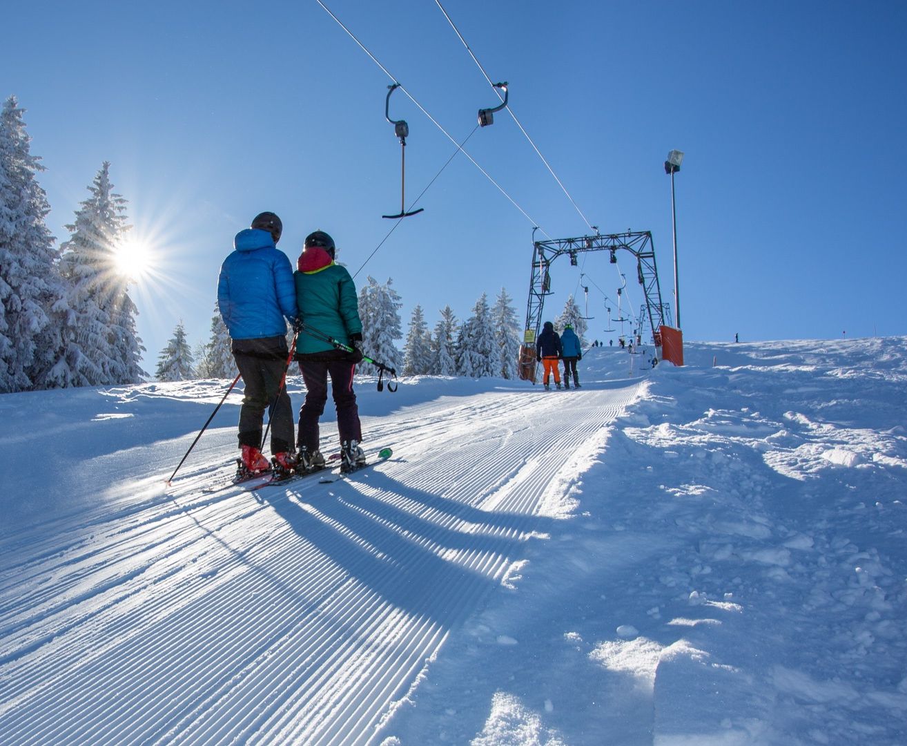 Zwei Skifahrer stehen auf einem verschneiten Hang unter einem strahlenden Sonnenschein und tragen Winterkleidung. Hinter ihnen ist eine Skiliftanlage zu sehen, an der Menschen Ski fahren.