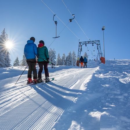 Zwei Skifahrer stehen auf einem verschneiten Hang unter einem strahlenden Sonnenschein und tragen Winterkleidung. Hinter ihnen ist eine Skiliftanlage zu sehen, an der Menschen Ski fahren.
