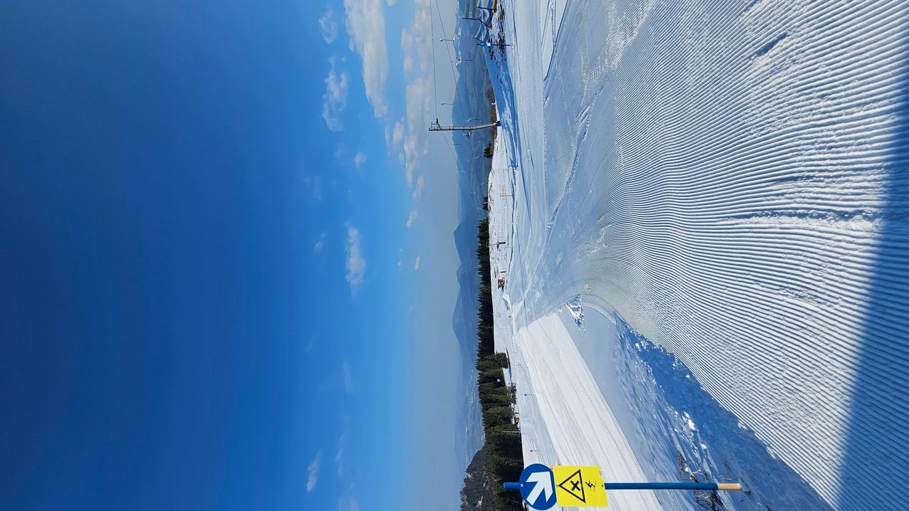 Ein schneebedeckter Berghang mit blauem Himmel und verstreuten Wolken. Ein gelbes Schild mit einem Skifahrer ist sichtbar. Bäume säumen die Seite des Hangs.