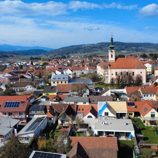 Luftaufnahme einer Stadt mit vielen Häusern, einige mit Solarpanels, unter einem blauen Himmel mit Wolken. Im Hintergrund gibt es Hügel und eine Kirche mit einem Turm.