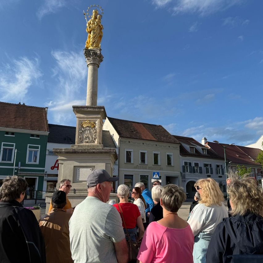 Eine Gruppe von Menschen steht um eine Statue auf dem Stadtplatz und blickt in einen blauen Himmel mit Wolken. Die Statue zeigt eine goldene Figur mit Krone und Zepter. Gebäude mit roten Dächern sind hinter der Statue sichtbar.