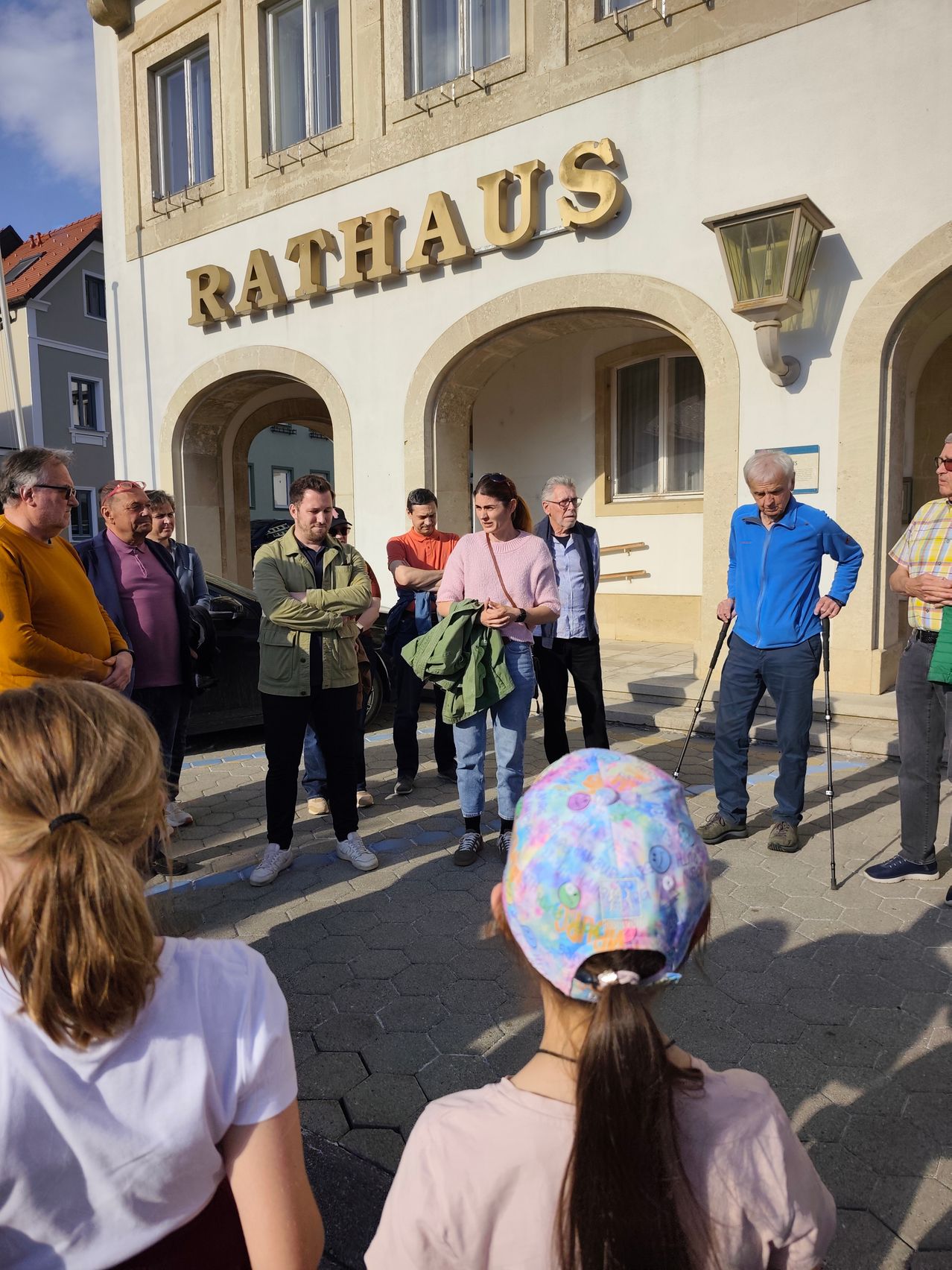 Eine Gruppe von Menschen versammelt sich vor einem Gebäude namens Rathaus. Einige tragen eine Brille und andere benutzen Stöcke. Sie befinden sich in einem sonnigen Außenbereich.