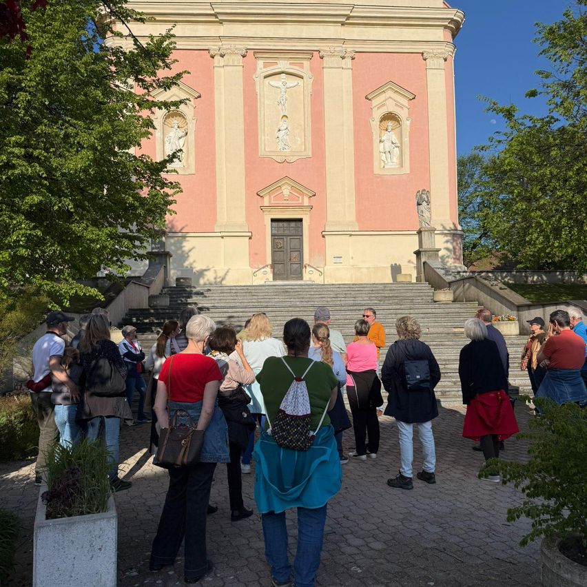 Eine Gruppe von Menschen steht vor einer Kirche mit rosa Fassade. Die Kirche hat Statuen und Pflanzen. Sie warten auf etwas.