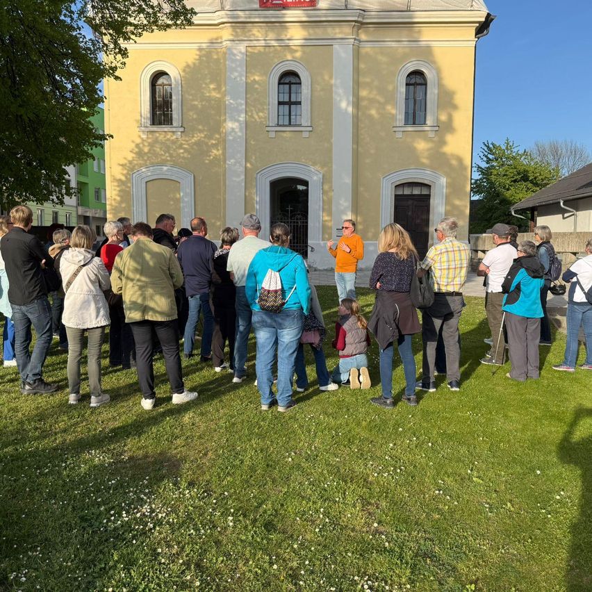 Eine Gruppe von Menschen versammelt sich auf einer Wiese vor einem gelben Gebäude. Einige lauschen einem Mann, der in der Nähe des Eingangs spricht.