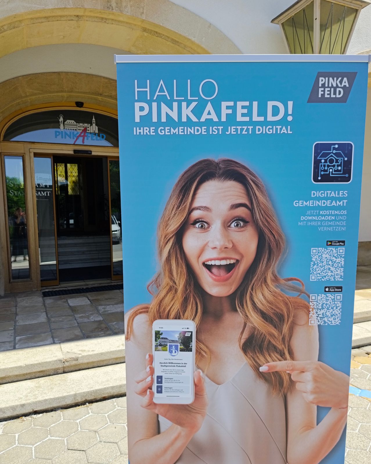 Werbung für Hello Pinkafeld mit einer Frau, die ein Handy hält und auf den Bildschirm zeigt. QR-Codes für App-Stores sind sichtbar.