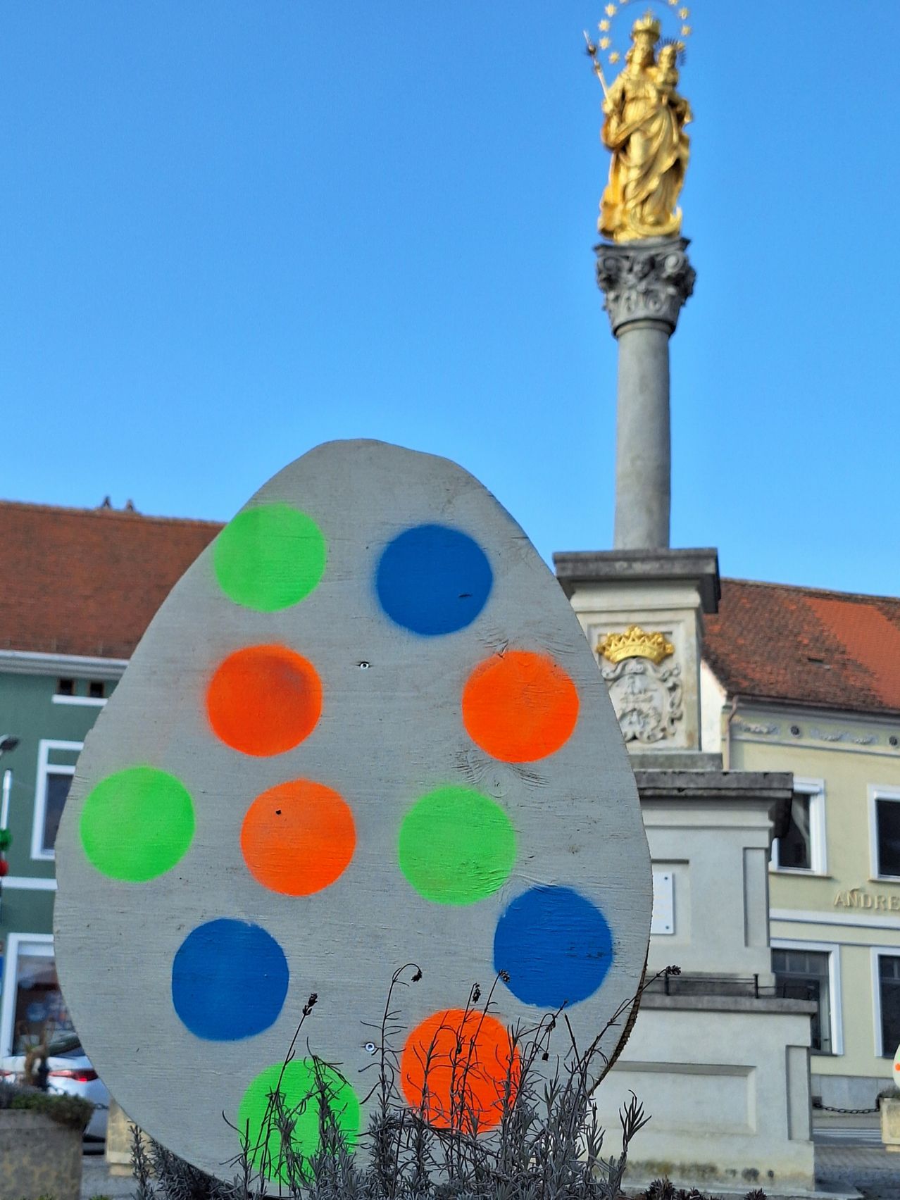 Ein farbenfrohes Holz-Osterei steht vor einem Denkmal mit einer goldenen Statue oben, vor einem Hintergrund von Gebäuden und einem klaren blauen Himmel.