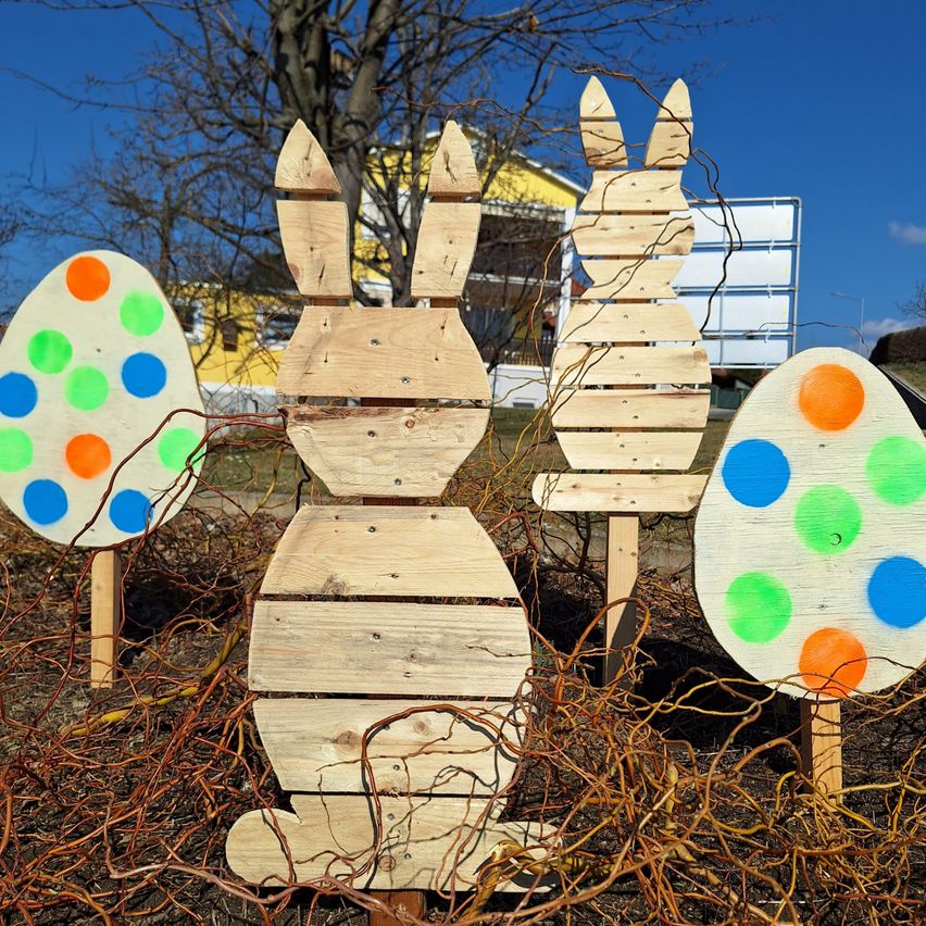 Drei hölzerne Osterdekorationen mit bunten Punkten stehen auf einem Erdboden mit Ästen. Dahinter befindet sich ein gelbes Gebäude mit einem klaren blauen Himmel darüber.