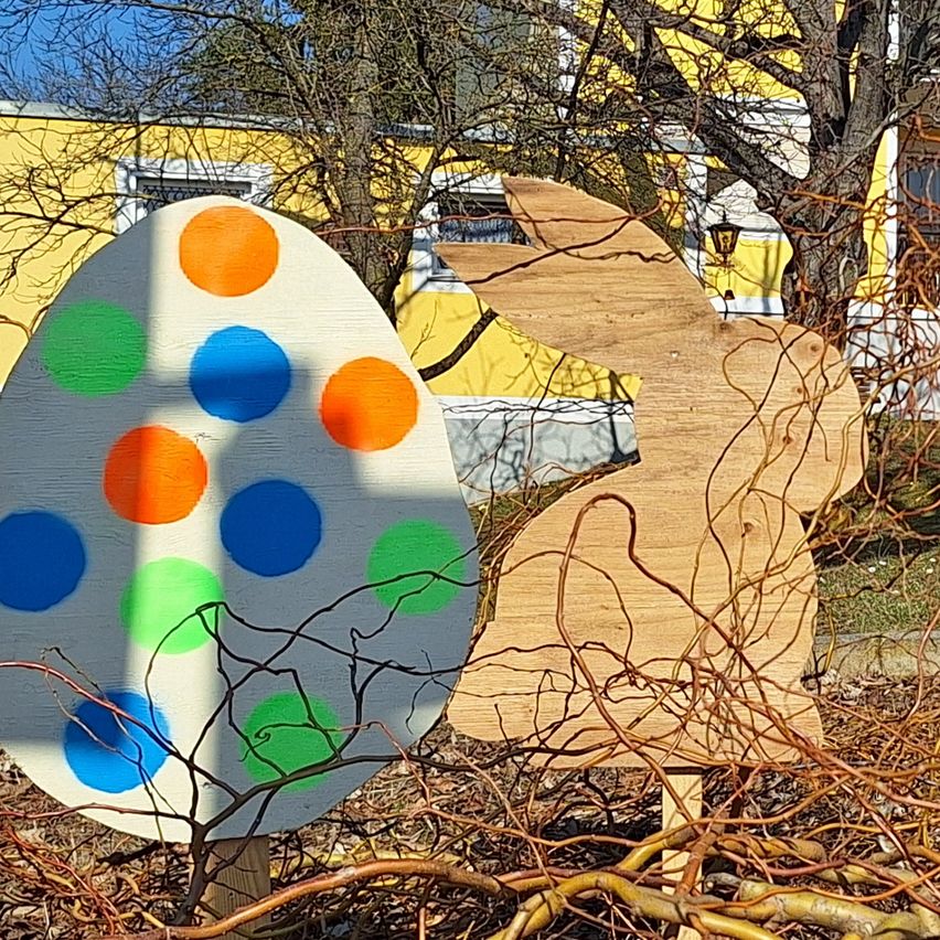 Ein hölzerne Ostereier mit bunten Punkten und ein Hase auf einem Busch vor einem gelben Haus.