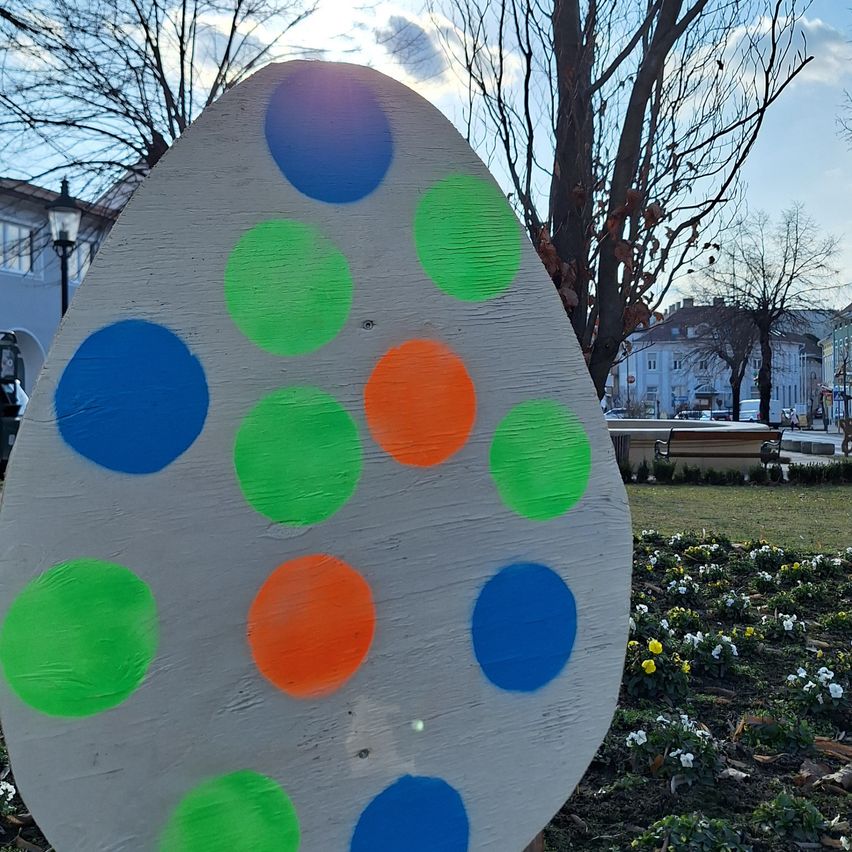Ein großes bemaltes Holzei mit grünen, blauen und orangefarbenen Punkten steht in einem Park mit einem Gebäude und einem Baum im Hintergrund.