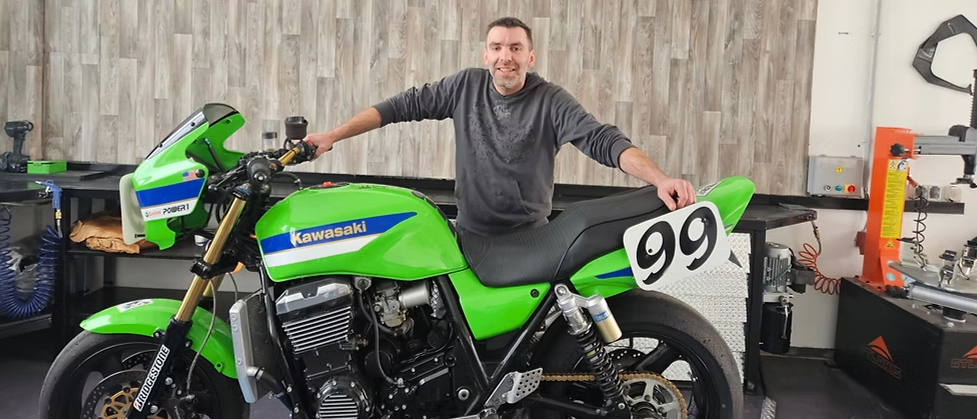 Ein Mann sitzt auf einer grünen Kawasaki-Motorrad mit der Nummer 99 vorne, lächelt und posiert für ein Foto in einer Garage.