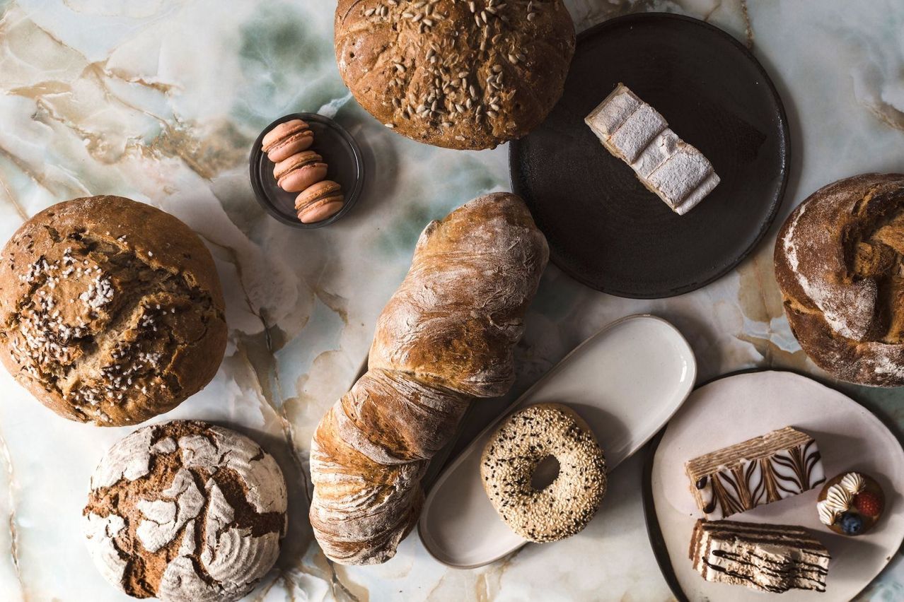 Auf einem Tisch sind verschiedene Brote und Gebäckstücke zu sehen. Ein Laib mit Samen liegt auf einem schwarzen Teller, während ein Croissant auf einem anderen steht. Ein weißer Teller mit Sesam-Bagel und ein weißer Teller mit schokoladenüberzogenen Desserts sind in der Nähe.