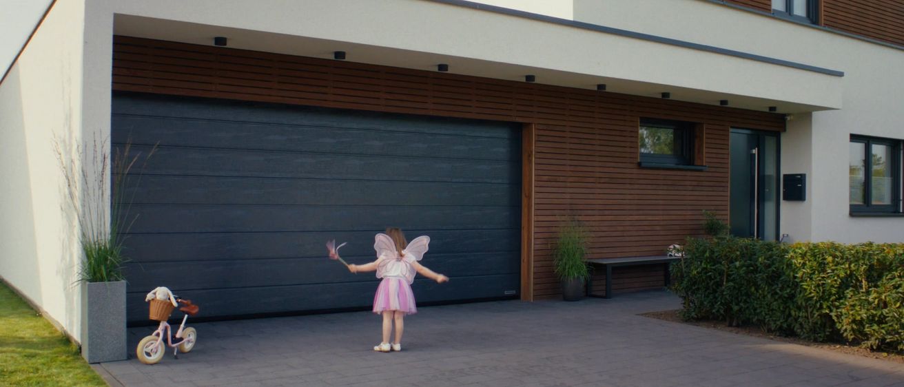 Ein junges Mädchen in einer Feen-Verkleidung steht vor einem modernen Haus und hält einen Zauberstab. Das Haus hat eine schwarze Garagentür und Holzwände.