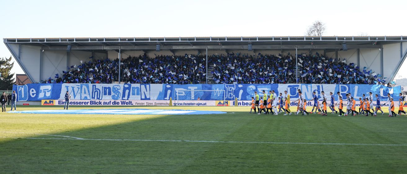 Ein Fußballspiel ist im Gange, Spieler laufen auf dem Feld. Viele Zuschauer füllen die Tribünen und halten blaue Fahnen hoch. Das Feld hat ein großes Banner mit Text und Logos.
