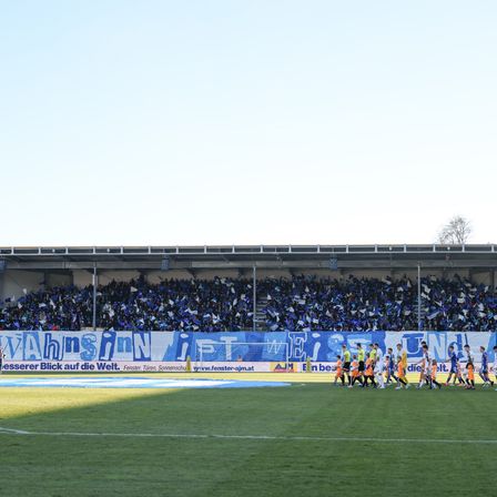 Fußballspieler in orangefarbenen Trikots laufen über ein Feld zum Tor. Zuschauer in blauen Trikots jubeln in den Rängen.