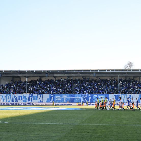Fußballspieler in orangefarbenen Trikots laufen über ein Feld zum Tor. Zuschauer in blauen Trikots jubeln in den Rängen.