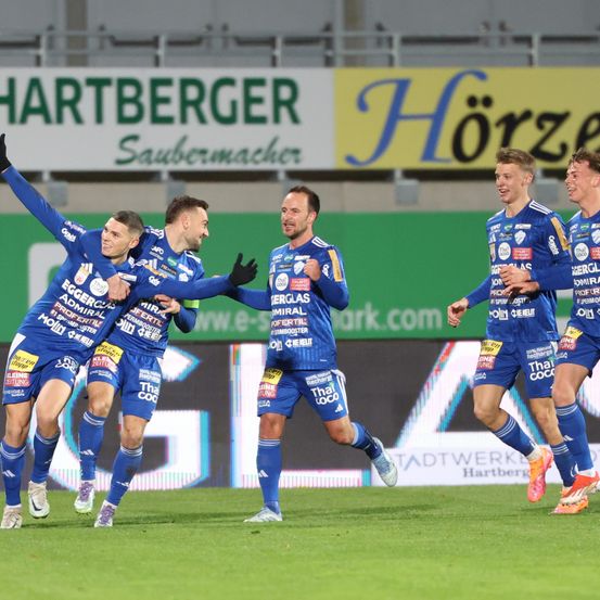 Fußballspieler in blauen Trikots feiern auf einem Feld mit grünen Bannern im Hintergrund. Das Wort 'Hartberger' ist auf einem weißen Banner zu sehen.