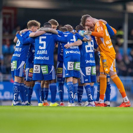 Eine Gruppe von Fußballspielern in blauen Trikots steht auf dem Feld eng beieinander. Der Spieler mit der Nummer 5 hält sich den Kopf, während die anderen ihn trösten. Ein gegnerischer Spieler in Orange steht in der Nähe.