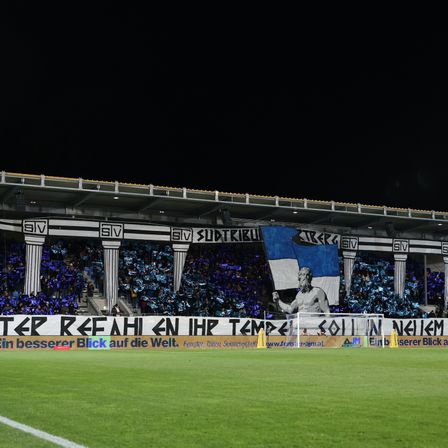 Fußballfans jubeln bei Nacht. Das Banner zeigt eine Statue und den Text 'Besserer Blick auf die Welt.' Das Stadion ist gefüllt mit Fans, und eine große Flagge ist auf der Brüstung angebracht.