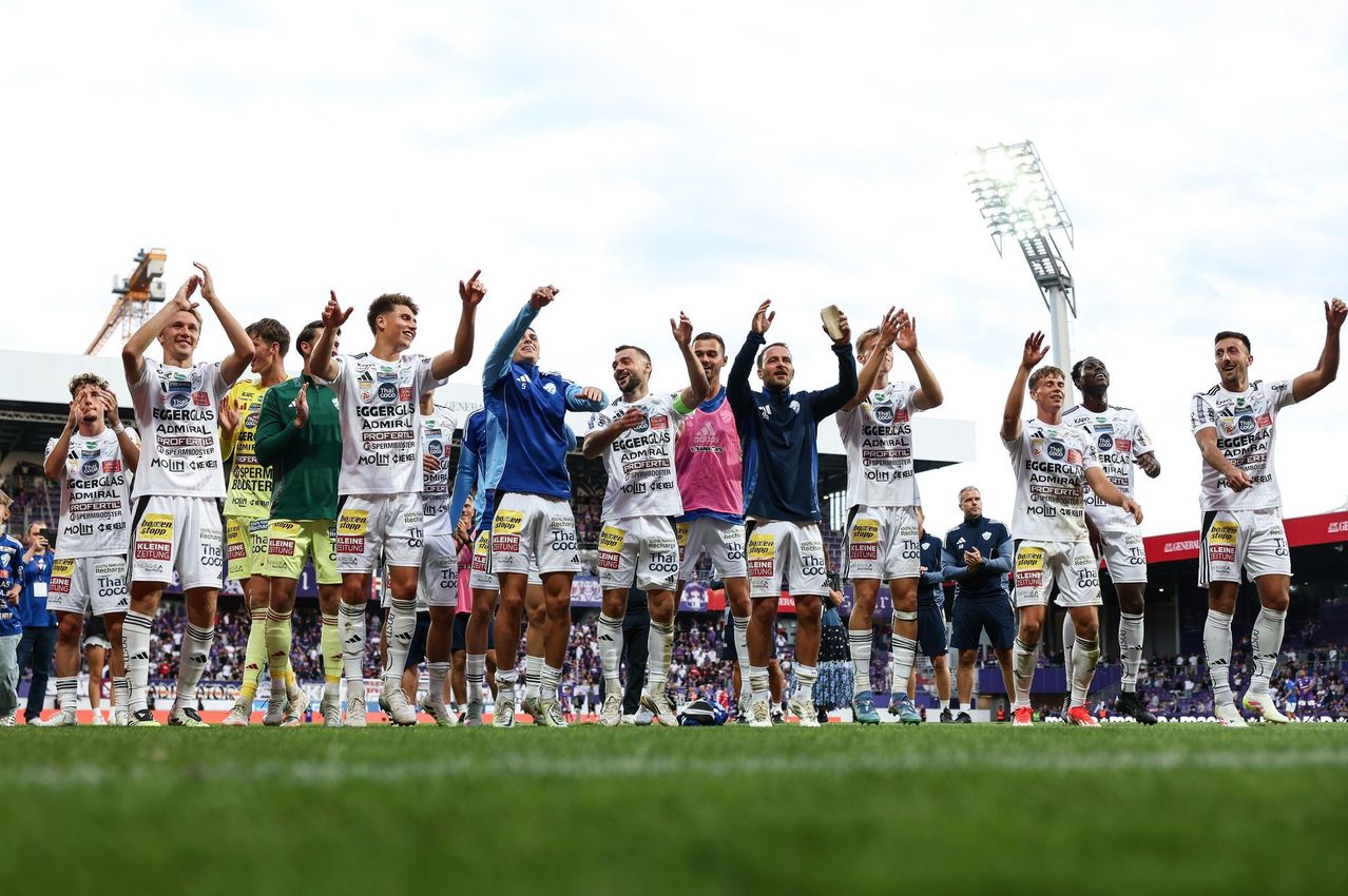 Eine Fußballmannschaft feiert auf einem Rasenfeld, während eine Menge zuschaut, unter einem teilweise bewölkten Himmel. Die Spieler tragen Trikots mit Werbung.