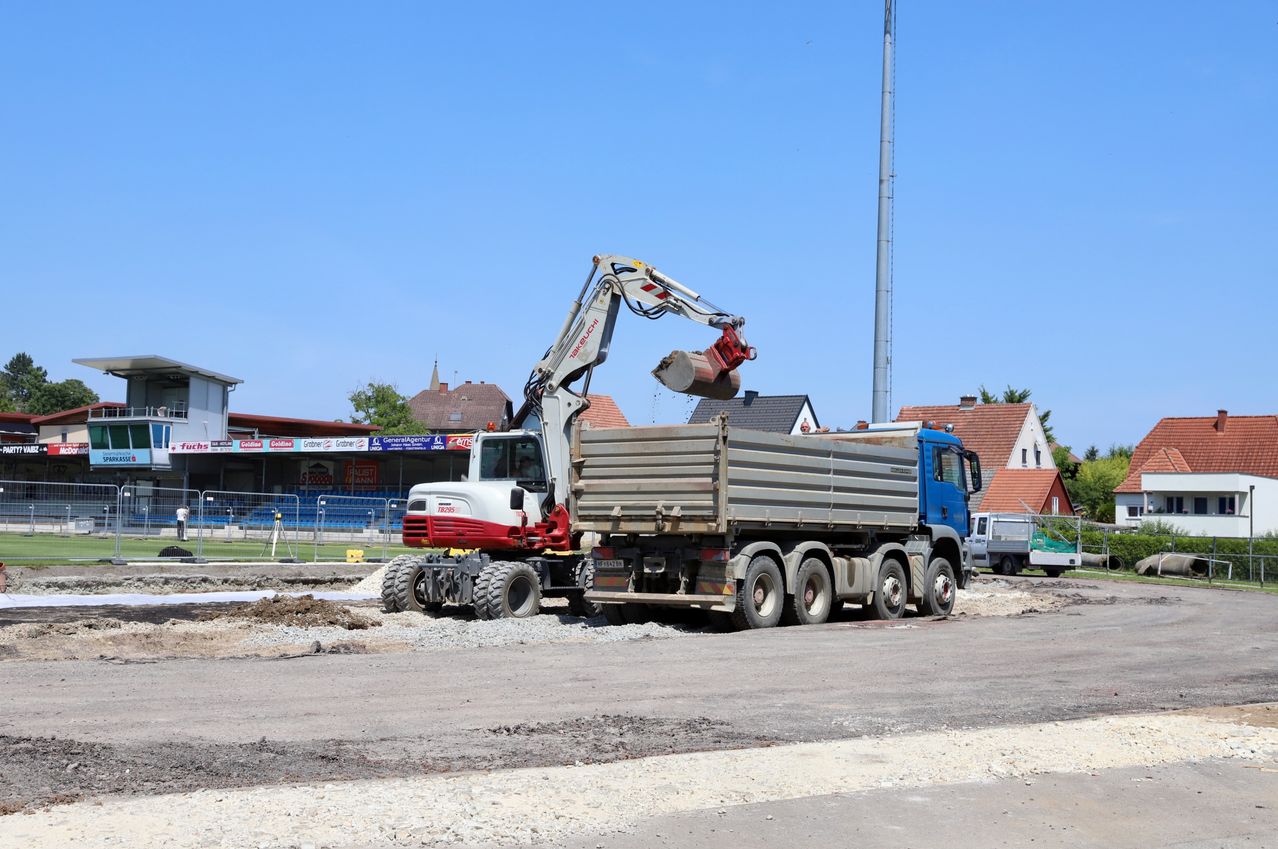 Eine Baustelle mit einem blauen und weißen Lastwagen und einem roten Kran an einem sonnigen Tag. In der Nähe befinden sich Häuser, ein Gebäude mit einem leeren Stadion und ein hoher Metallturm.