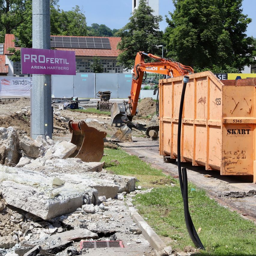 Eine Baustelle mit einem Schild für Profertil Arena Hartberg, einem Bagger, einem Container, Schutt und einem Straßenlaterne.