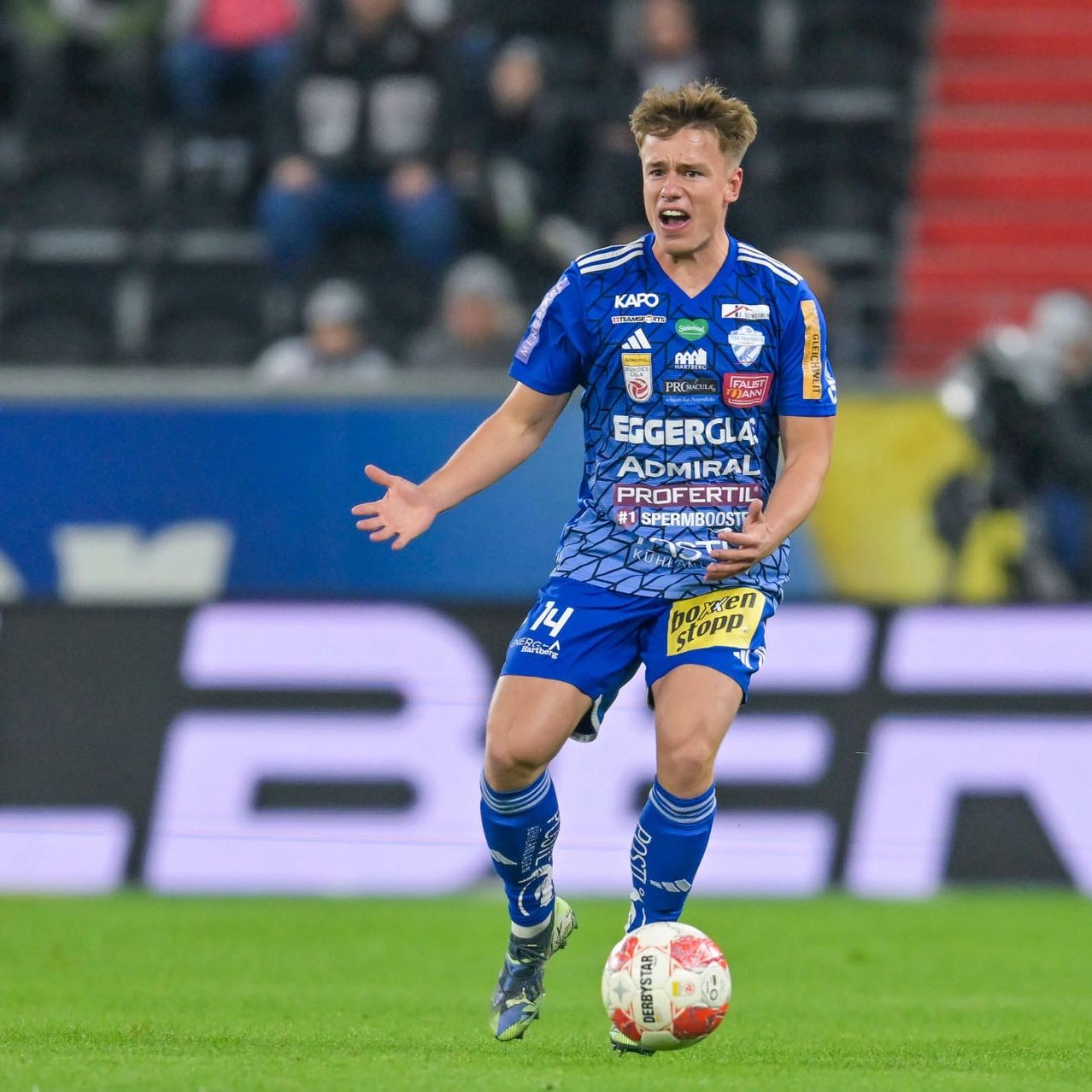 Ein Fußballspieler in blauem Trikot mit vielen Werbeaufklebern läuft mit einem Fußball auf dem Feld.