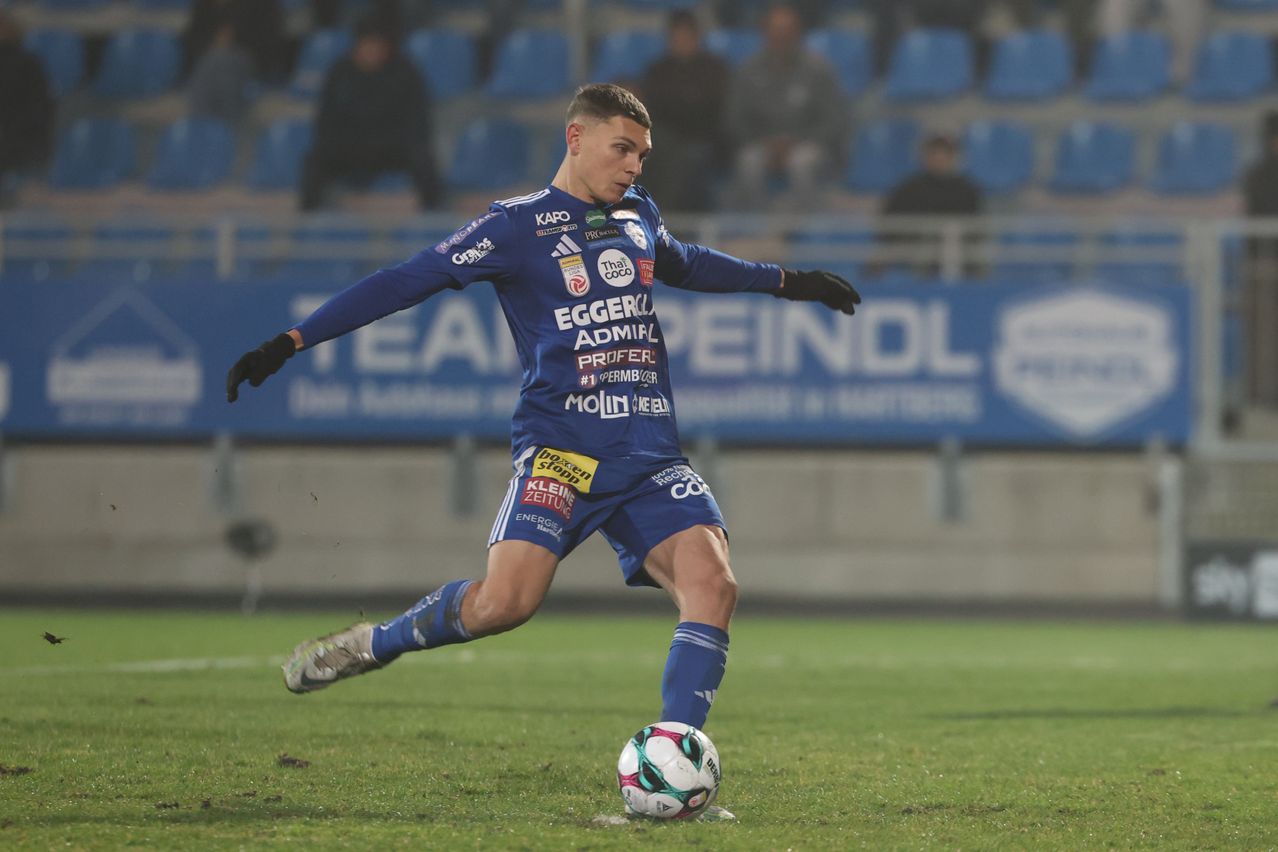 Ein Fußballspieler in blauer Uniform tritt während eines Spiels einen Ball, mit verschwommenem Hintergrund von blauen Sitzen und Menschen.