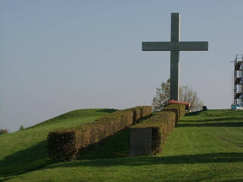 Ein großes Kreuz steht auf einem Hügel, umgeben von einer Hecke. Hinter der Hecke ist eine Betonstruktur zu sehen. Der Hügel ist mit saftigem grünem Gras bedeckt. In der Ferne ist ein Turm zu erkennen, und hinter dem Kreuz befindet sich ein Baum.