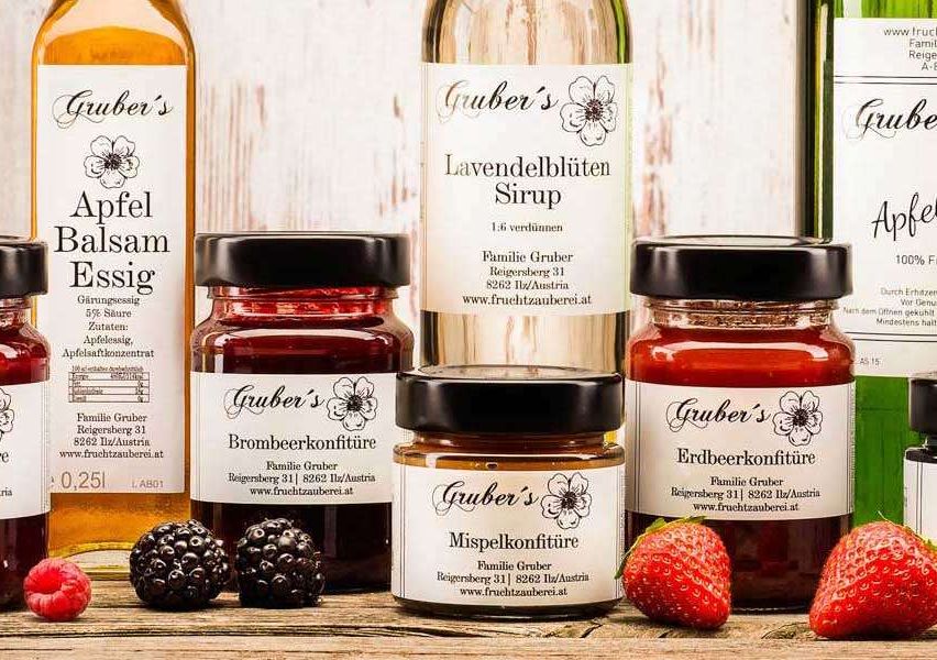 Auf einem Holztisch sind verschiedene Glasgläser und Flaschen von Grueber's Fruchtkonserven und Sirupen ausgestellt. Die Produkte umfassen Himbeermarmelade, Brombeermarmelade, Erdbeermarmelade, Apfelbalsamico, Lavendelsirup und Himbeersirup. Die Gläser und Flaschen sind mit dem Markennamen 'Grueber's' beschriftet und enthalten eine Mischung aus Obst und Sirup.
