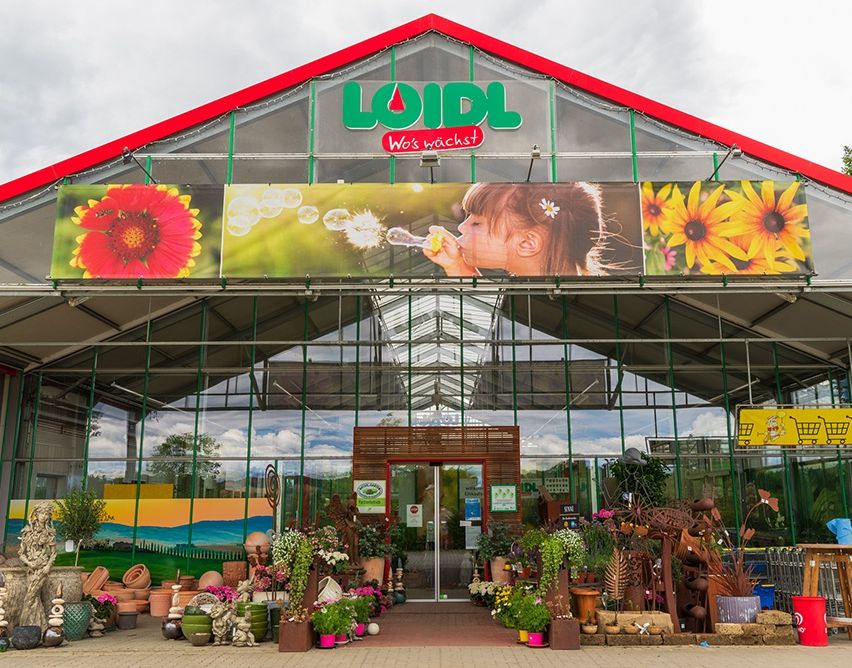 Ein Bild eines Gartencenters mit einem Mädchen, das draußen Seifenblasen bläst. Das Geschäft hat ein rotes Dach, grüne Wände und große Glasfenster. Das Banner über dem Eingang zeigt ein Mädchen, das Seifenblasen bläst und Blumen.