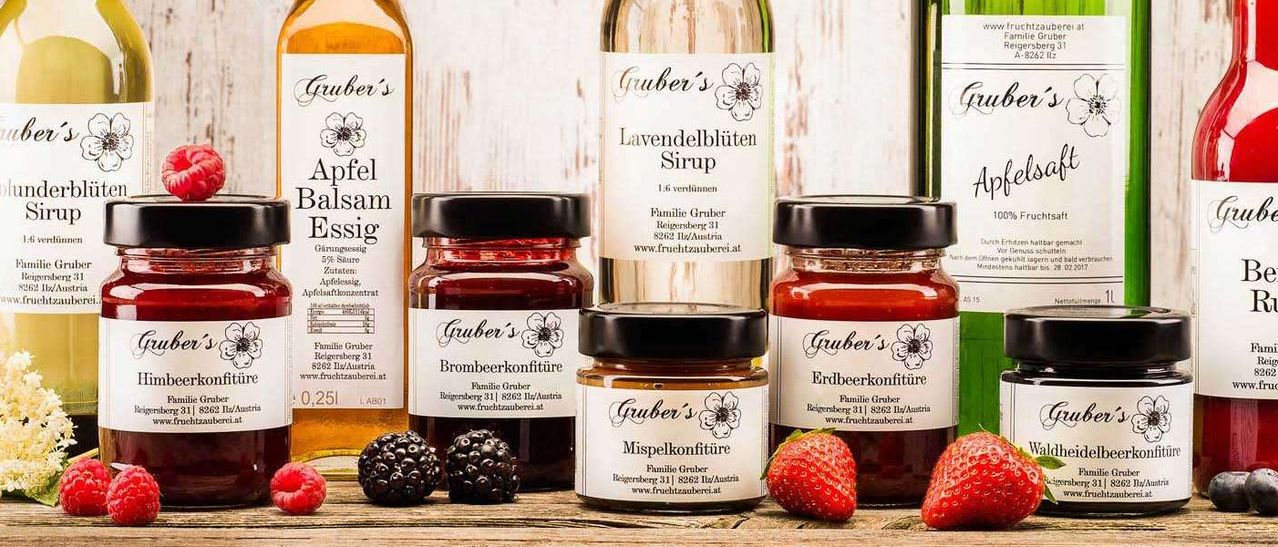 Eine Sammlung von Gruber's Fruchtkonserven und Sirupen, einschließlich Apfel-Balsamessig, Brombeer-, Himbeer- und Lavendelsirup, auf einer Holzoberfläche ausgestellt.