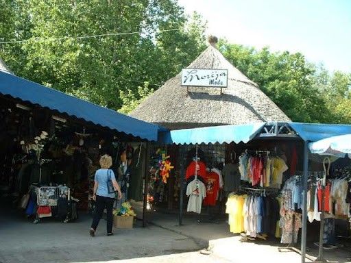 Ein Outdoor-Kleidungmarkt mit einem Strohdach, der eine Vielzahl von Hemden unter blauen Markisen zur Schau stellt. Eine Frau in einem blauen Shirt und schwarzen Hosen geht an den Ständen vorbei.