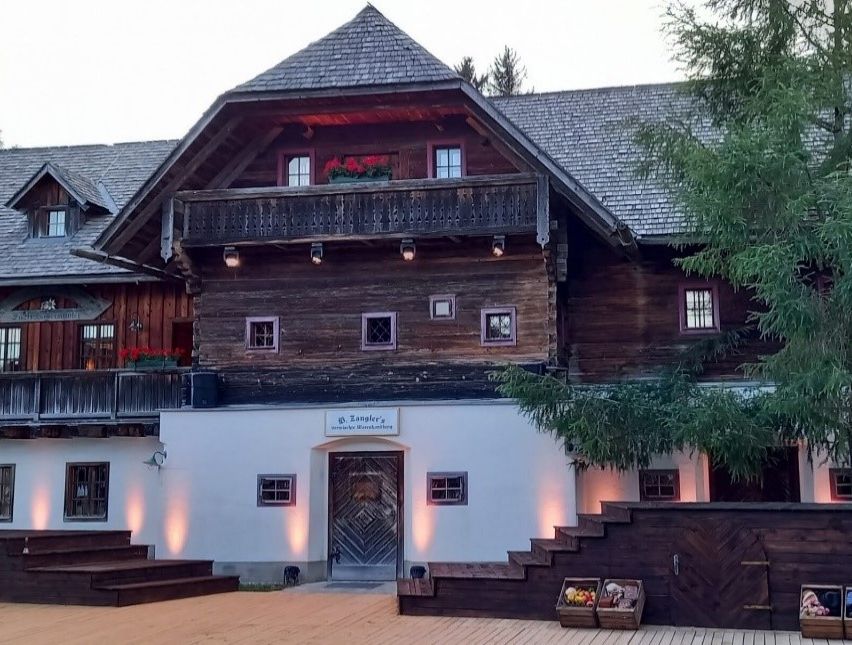 Ein traditionelles Holzhaus mit Balkon und roten Blumen im Fensterkasten. Das Haus ist gut beleuchtet und von Bäumen umgeben.