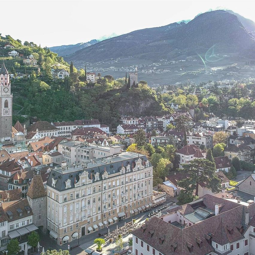 Luftaufnahme einer europäischen Stadt mit einem großen Gebäude im Vordergrund, umgeben von Bäumen und Bergen.