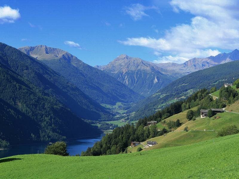 Ein friedliches Tal, umgeben von Bergen, mit einem ruhigen See und verstreuten Häusern unter einem teilweise bewölkten Himmel.