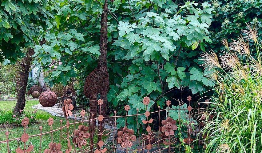 Ein Garten mit einem dekorativen Metallzaun mit Blumendesigns, einer Giraffenskulptur und verschiedenen Pflanzen im Hintergrund.