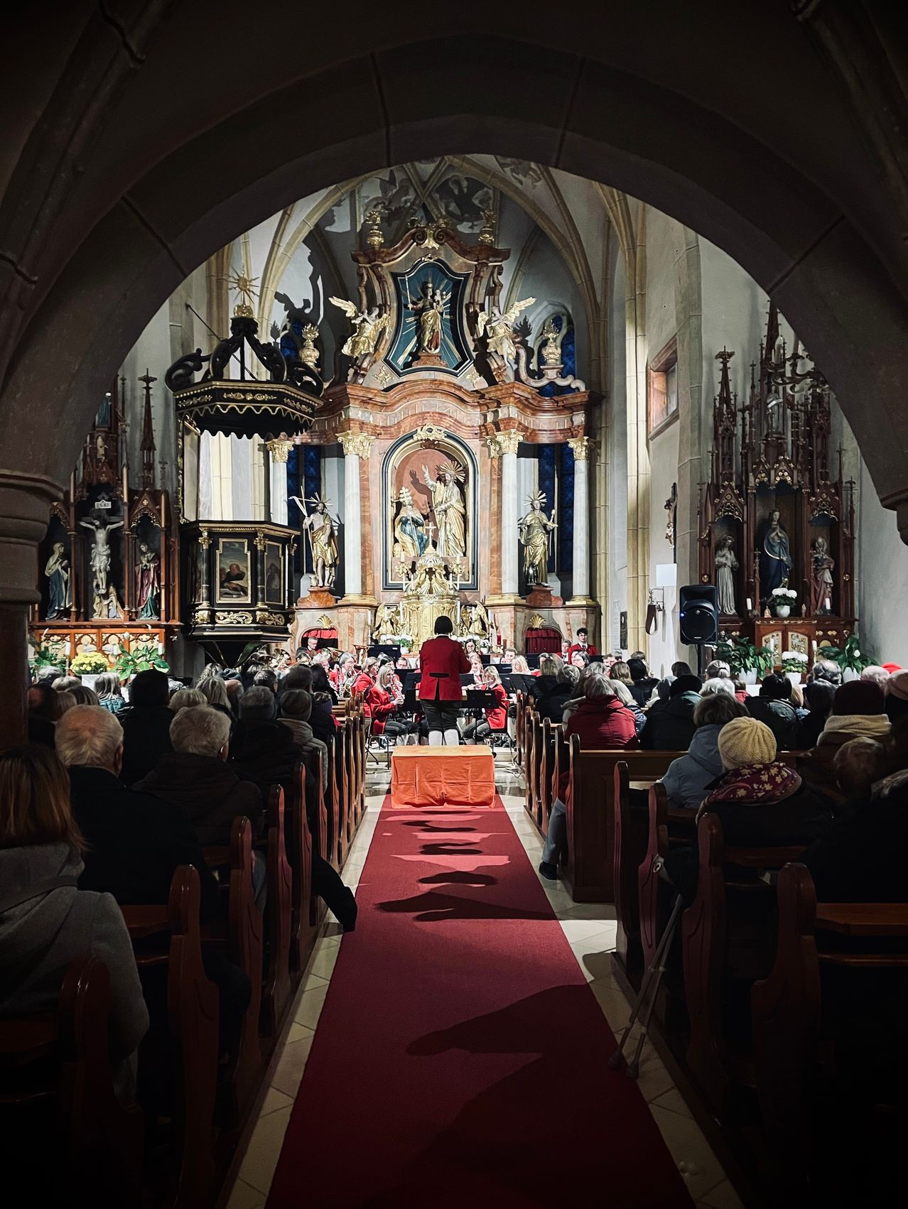 Ein Chor tritt in einer Kirche mit einem kunstvollen Altar und religiösen Statuen auf. Das Publikum sitzt auf hölzernen Kirchenbänken, einige tragen Hüte. Ein roter Teppich führt zur Bühne.