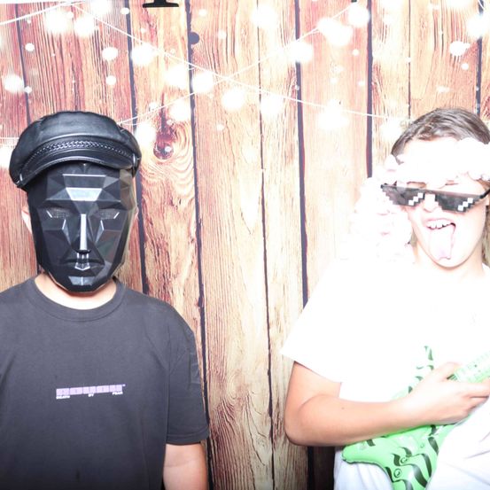 Zwei junge Personen posieren für ein Foto vor einem hölzernen Hintergrund. Einer trägt eine schwarze Maske, der andere ein weißes T-Shirt und hält eine grüne Gitarre.