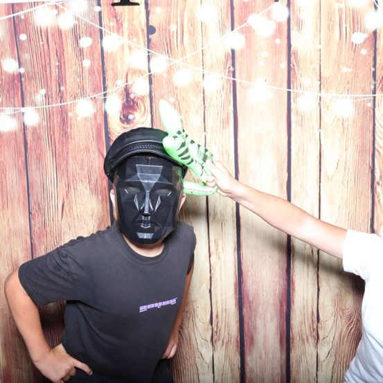 Eine Person in einer schwarzen Maske und einem schwarzen T-Shirt steht vor einer Holzwand mit Lichterketten. Eine andere Person in einem weißen T-Shirt hält ein grünes Spielzeug vor das Gesicht der maskierten Person.