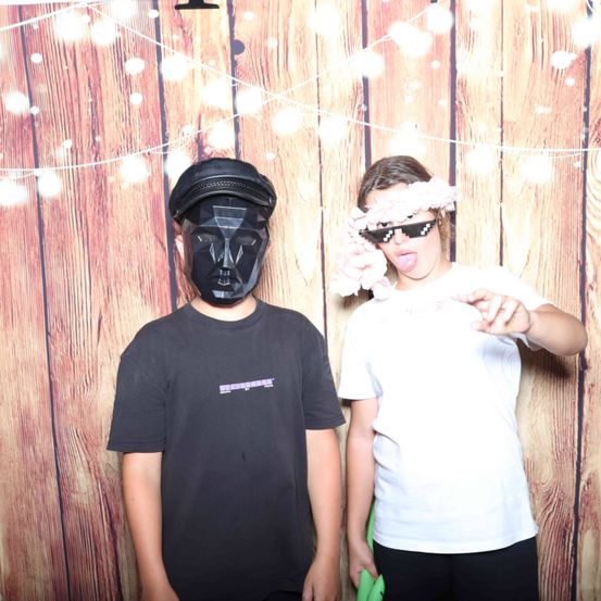 Zwei Personen posieren vor einem hölzernen Hintergrund. Die Person auf der linken Seite trägt eine schwarze Maske, während die Person auf der rechten Seite eine pinke Blumengirlande und eine Sonnenbrille trägt.