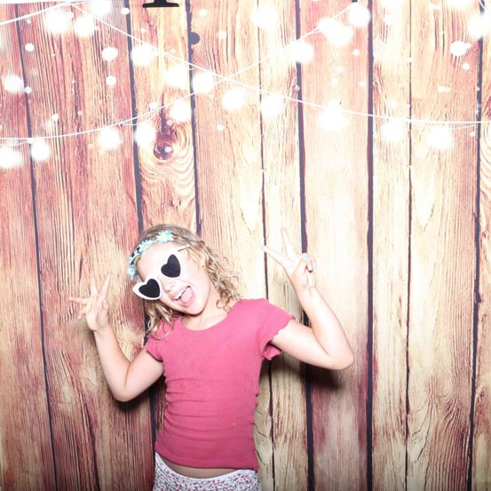 Ein junges Mädchen steht vor einem hölzernen Hintergrund, trägt ein rosa T-Shirt und herzförmige Sonnenbrille, lächelt und macht ein Friedenszeichen.