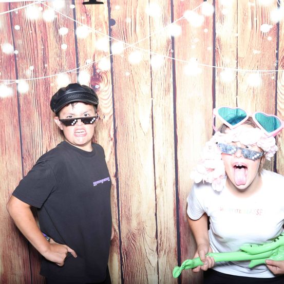 Zwei Kinder stehen vor einer Holzwand. Der Junge trägt einen schwarzen Hut und Sonnenbrille, während das Mädchen rosa Haare, herzförmige Brillen und eine grüne Spielzeuggitarre hat. Sie lächeln beide für die Kamera.