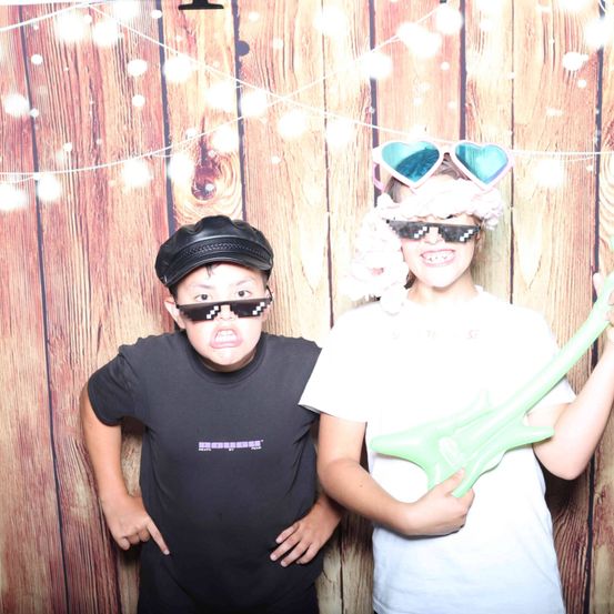 Zwei Kinder stehen in einem Fotoautomaten mit einem hölzernen Hintergrund. Ein Junge trägt einen schwarzen Hut und eine Sonnenbrille, während ein Mädchen herzförmige Brillen und eine rosa Perücke trägt. Beide halten Spielzeuggitarren.