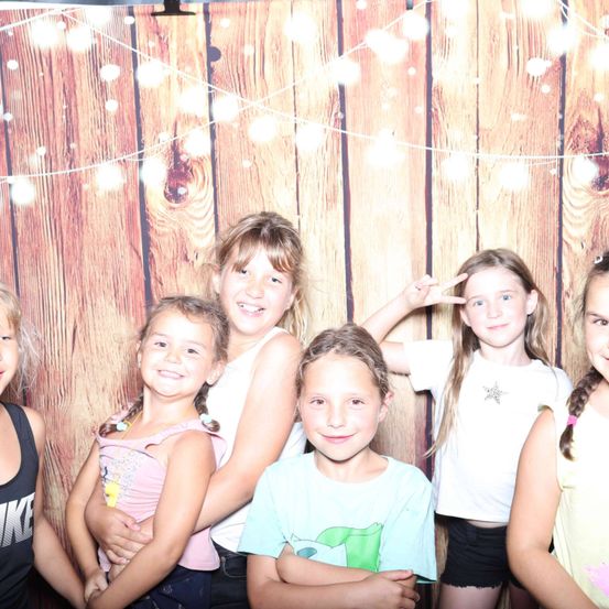 Sechs junge Mädchen posieren zusammen vor einem hölzernen Hintergrund mit Lichtern und Dekorationen. Sie lächeln und scheinen Spaß zu haben.