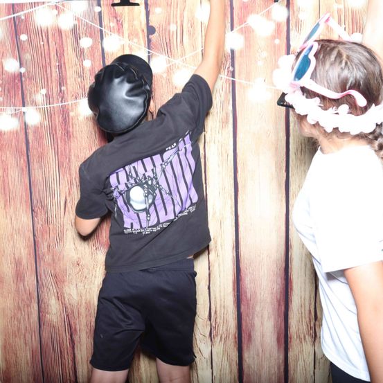 Zwei Kinder stehen vor einem hölzernen Hintergrund. Ein Kind trägt einen schwarzen Helm und ein Shirt mit einem lila Design. Das andere Kind trägt ein weißes T-Shirt und eine Kopfbedeckung mit rosa Blumen.