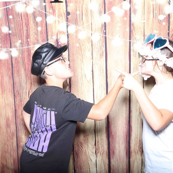 Zwei Kinder stehen in einem Fotokabine. Der Junge trägt einen schwarzen Hut und Brille und zeigt. Das Mädchen trägt eine Brille und eine Blumenkrone und zeigt auch. Dahinter ist eine hölzerne Wand mit Lichtern.