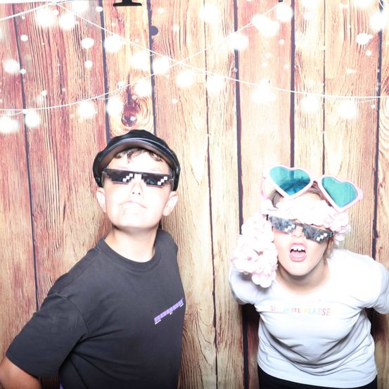 Zwei Kinder stehen vor einem hölzernen Hintergrund und tragen Sonnenbrillen. Der Junge hat einen schwarzen Hut und ein schwarzes T-Shirt. Das Mädchen hat herzförmige Sonnenbrillen, rosa Haare und ein weißes T-Shirt.