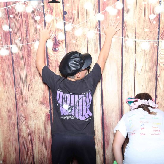 Ein Junge steht mit erhobenen Händen vor einem hölzernen Hintergrund, trägt einen schwarzen Hut und ein schwarzes T-Shirt mit einem grafischen Design. Neben ihm steht ein Mädchen mit einer geblümten Haarspange, das mit dem Rücken zur Kamera steht.