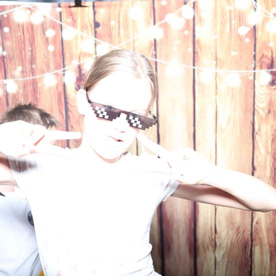 Ein junges Mädchen mit pixeligen Brillen posiert mit einem Friedenszeichen vor einem hölzernen Hintergrund. Eine andere Person ist teilweise hinter ihr sichtbar.