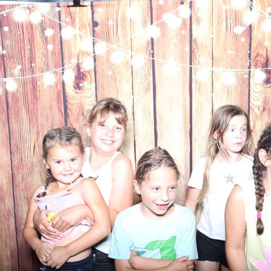 Fünf Mädchen posieren für ein Foto vor einem hölzernen Hintergrund mit dekorativen Lichtern. Sie lächeln und scheinen sich zu amüsieren.